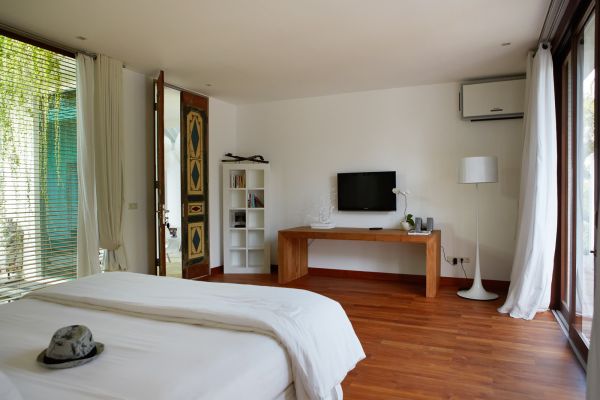 Spacious tropical bedroom with white linens on king bed, wooden desk and TV, open sliding doors to lush greenery, minimalist decor.