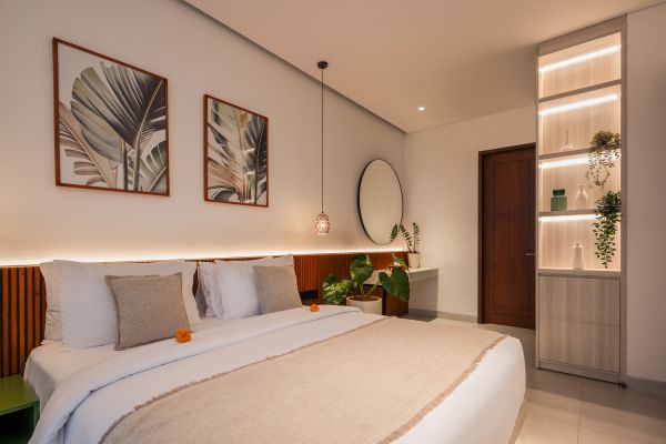 Cozy Balinese-style hotel bedroom with white linens, wooden headboard, tropical plants, and leaf wall art.