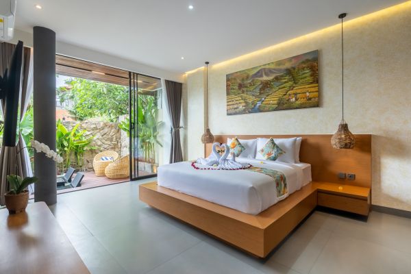 Spacious Balinese villa bedroom with wooden king bed, floral accents, tropical plants, and glass doors opening to a lush garden view. - Bali Villas