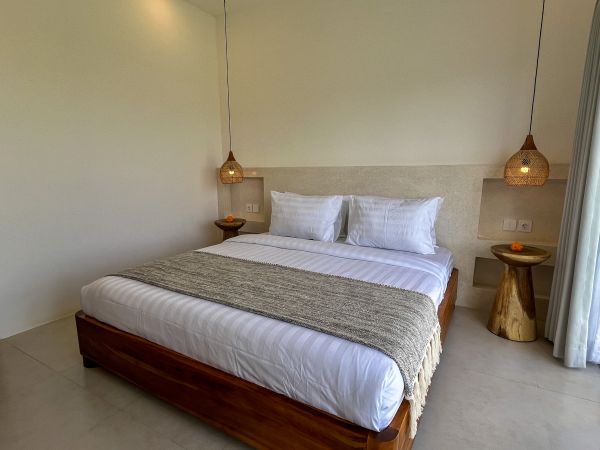Cozy tropical bedroom with wooden bed, white linens, gray throw blanket, rattan pendant lamps, and side tables beside sheer-curtained windows.