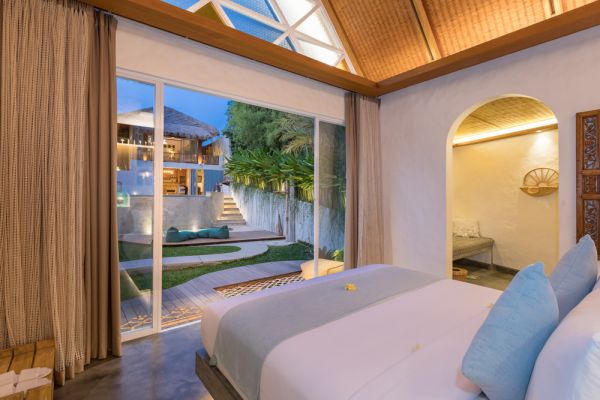 Serene tropical bedroom with wooden beamed ceiling, open sliding doors to lush garden and pool at dusk.