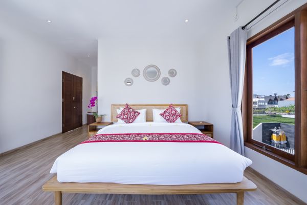 Spacious Balinese bedroom with wooden bed, pink floral pillows, woven wall decor, and tropical garden view through large windows.