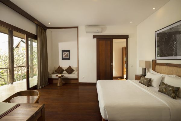 Spacious Bali villa bedroom with wooden floors, king bed, desk, lounge seating, and tropical garden views through large glass doors. - Bali Villas