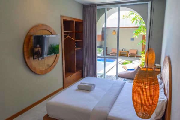 Cozy tropical bedroom with white bed, wooden wardrobe and mounted TV, sliding doors to lush courtyard pool. - Bali Villas