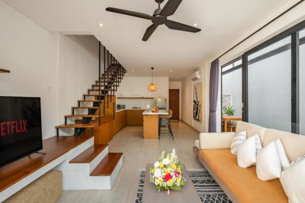 Modern open-plan living room with wooden staircase, kitchen island, beige sofa, floral coffee table, and Netflix on wall-mounted TV in a bright tropical villa. - Bali Villas