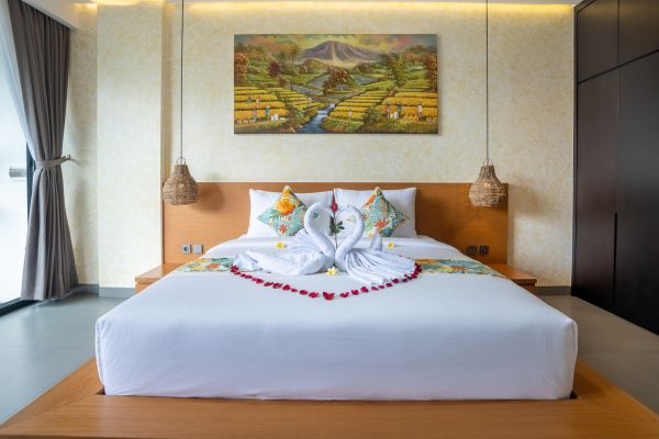 Luxurious Balinese bedroom with heart-shaped rose petals and towel swans on a king bed, framed by rice terrace artwork and sheer curtains.