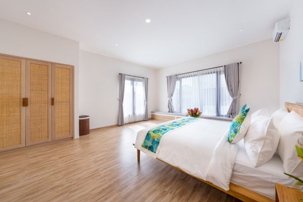 Spacious tropical bedroom with wooden bed, bamboo wardrobe, large curtained windows, and green accents on light wood floors.