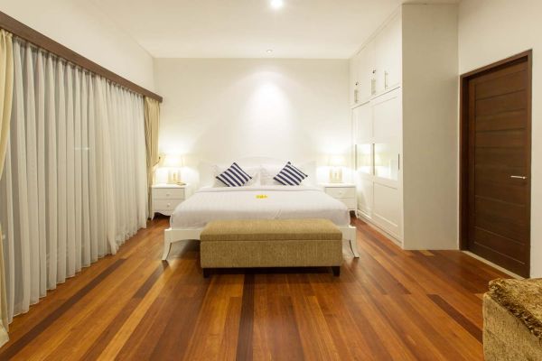 Serene Bali-style bedroom with white king bed, sheer curtains, wooden floors, and bedside lamps.