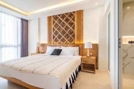 Modern tropical bedroom with intricate wooden lattice headboard, king bed with white linens, bedside lamps, large window, and adjacent marble bathroom.
