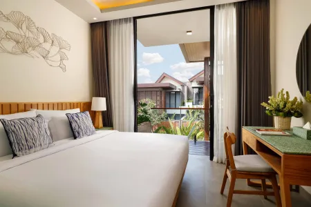 Spacious modern tropical hotel room with king bed, wooden desk, and open balcony overlooking lush greenery. - Bali Villas