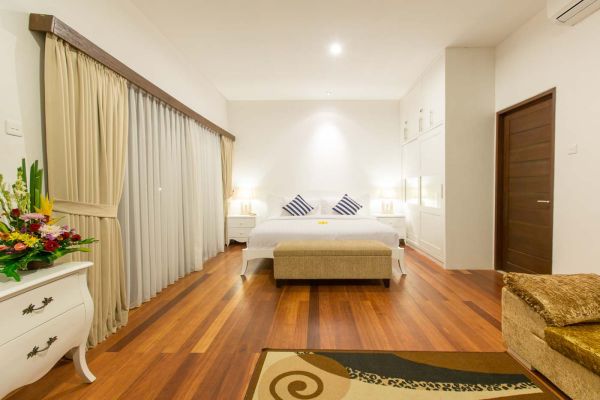 Spacious luxury Bali villa bedroom with king bed, beige curtains, wooden floors, bedside lamps, and fresh floral vase.
