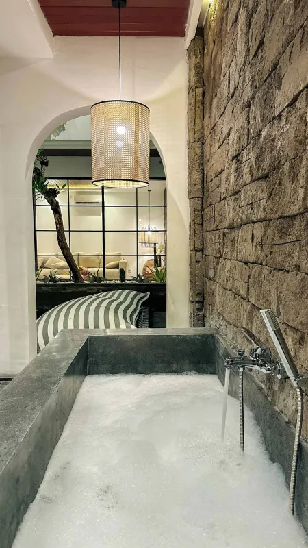 Modern rustic bathroom with a bubbling concrete soaking tub, exposed brick walls, rattan pendant light, and arched doorway to a plant-adorned bedroom.