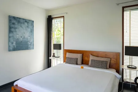 Modern minimalist bedroom with white bedding on a wooden headboard bed, abstract blue wall art, potted plant, and window views of greenery.
