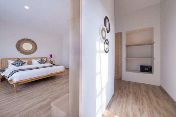 Modern Balinese-style bedroom with wooden bed, batik pillows, rattan mirrors, and built-in safe on light wood floors.