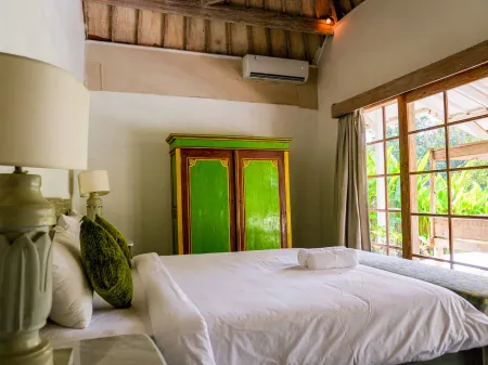 Cozy tropical bedroom with white bed, green armoire, potted plants, and lush green views through wooden windows.