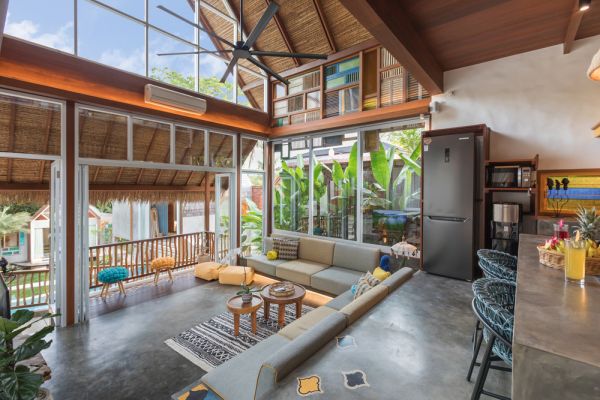 Spacious tropical Balinese villa interior with high wooden-beamed ceiling, large glass windows overlooking lush garden, plush sectional sofa, and modern kitchen bar. - Bali Villas
