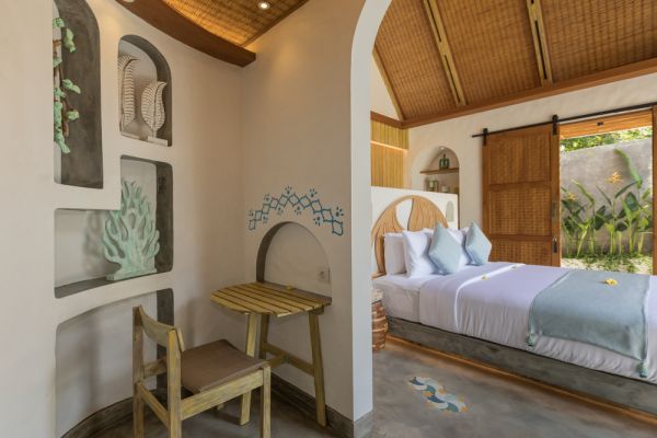 Cozy Balinese bedroom interior with arched niches, seashell decor, wooden beams, and open sliding door to tropical greenery.