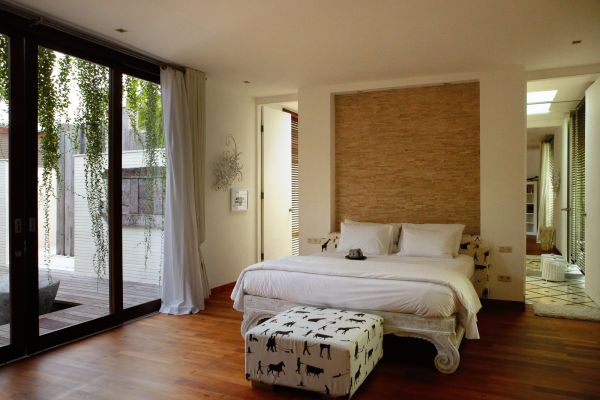 Modern tropical bedroom with white bed, black-and-white spotted ottoman, wooden floors, and large sliding doors opening to a lush balcony.