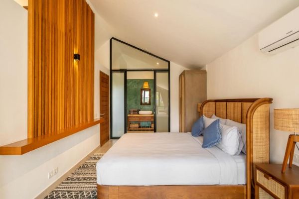 Modern tropical bedroom with wooden paneling, king bed with white linens, rattan lamps, and glass door to en-suite bathroom.