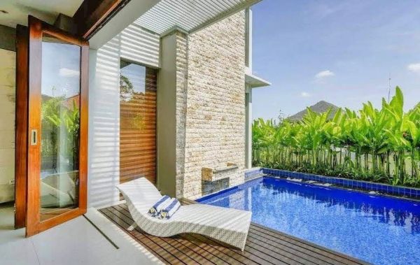 Modern outdoor infinity pool with white lounger chair, tropical plants, and wooden deck in a luxury Bali villa. - Bali Villas