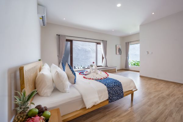 Luxurious tropical bedroom with rattan-framed king bed, swan towels, fresh fruits, and large windows overlooking greenery.
