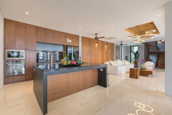 Modern open-plan kitchen and living area with wooden cabinets, black island countertop, white sofa, tropical plants, and ambient ceiling lights in a luxurious contemporary home. - Bali Villas