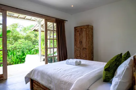 Serene tropical bedroom with open wooden doors to lush green garden, white linens on wooden bed, and antique wardrobe.