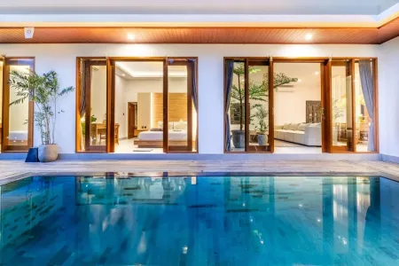 Luxurious open-plan Balinese villa bedroom with sliding doors overlooking a serene infinity pool, wooden accents, and tropical plants at dusk.