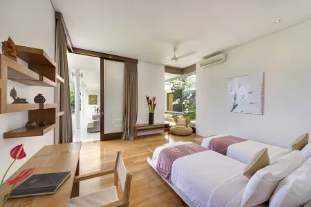 Serene Balinese villa bedroom with twin beds, wooden desk, tropical plants, and sliding doors to lush garden views.