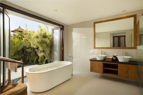 Luxurious Balinese-style bathroom with freestanding white bathtub filled with flower petals, wooden double vanity, and open doors revealing tropical garden view. - Bali Villas
