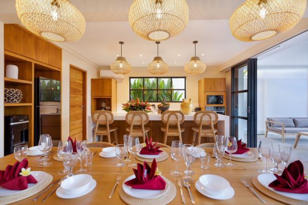 Elegant open-plan tropical kitchen with wooden cabinets, bamboo pendant lights, set dining table, and sliding glass doors to outdoor patio. - Bali Villas