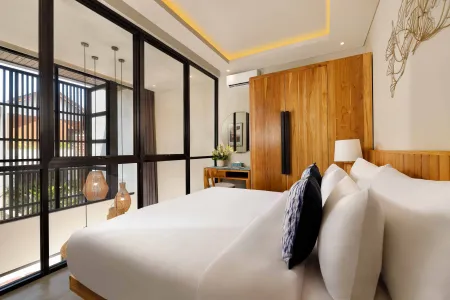 Modern tropical bedroom in Bali villa with white bed, wooden wardrobe, large black-framed windows overlooking lush greenery, and warm ambient lighting.