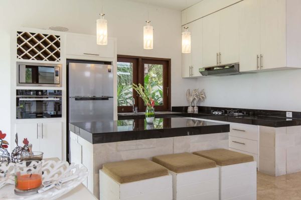 Modern tropical kitchen with white cabinets, black granite island bar stools, stainless steel appliances, and lush plant-filled windows. - Bali Villas