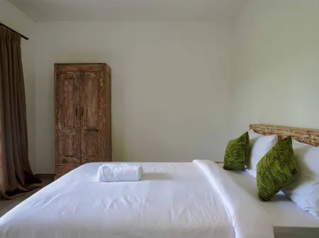 Cozy tropical bedroom with white bed, green pillows, antique wooden wardrobe, and light green walls.