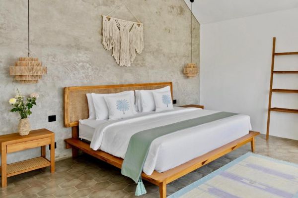 Serene Balinese bedroom with wooden platform bed, white linens accented by green throw, palm-embroidered pillows, macrame wall hanging, rattan lamps, and ladder shelf on textured concrete walls. - Bali Villas