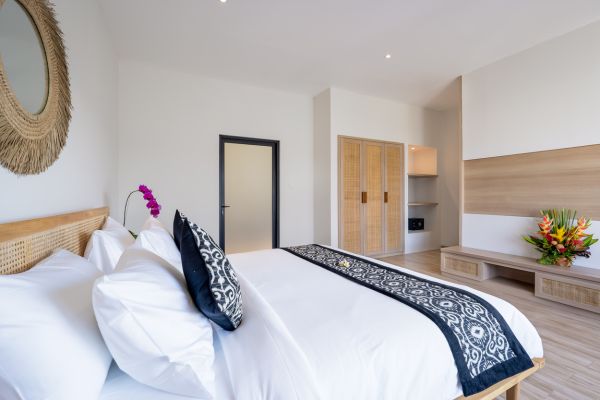 Spacious Balinese-style bedroom with wooden rattan bed, white linens, purple orchids, tropical flower vase, and round woven mirror on white walls.