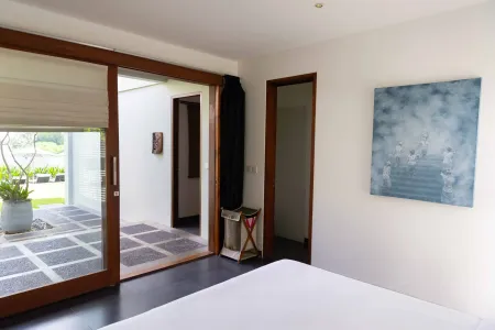 Modern bedroom interior with open sliding glass doors revealing a tropical courtyard, abstract blue wall art, and minimalist decor.