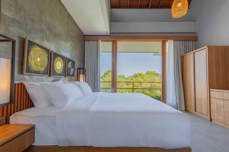 Spacious Balinese-style bedroom with king bed, abstract gold art, wooden accents, and lush tropical garden view through open sliding doors.