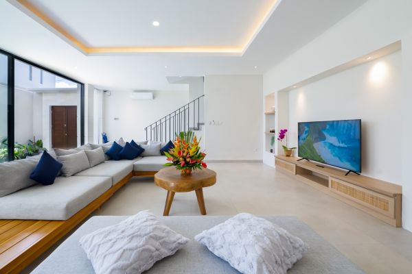 Modern tropical living room with L-shaped gray sofa, wooden coffee table topped with flowers, large glass windows, potted plants, and wall-mounted TV on bamboo stand. - Bali Villas