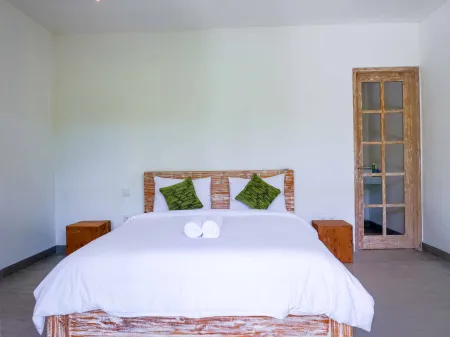Serene tropical bedroom with white bed on rustic wooden frame, green pillows, folded towels, wooden nightstands, and glass-paneled door.