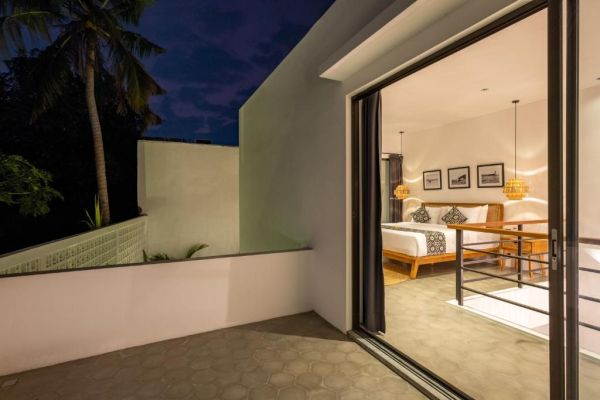 Nighttime view of a modern Bali villa bedroom with wooden bed and pendant lights, open sliding doors to a tiled balcony overlooking palm trees and lush greenery.