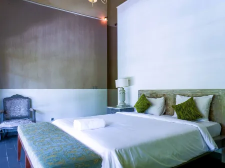 Cozy hotel bedroom featuring a white bed with green pillows, ornate upholstered armchair, bedside lamp, and neutral walls.