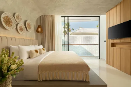 Luxurious modern bedroom with beige bedding, woven wall baskets, fresh lilies, mounted TV, and sliding doors to a tropical palm tree view. - Bali Villas