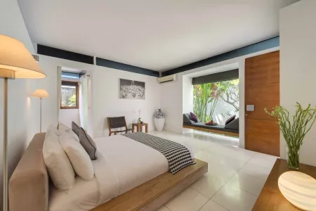Spacious Bali villa bedroom with wooden platform bed, tropical window view, potted plants, and soft lamp lighting.
