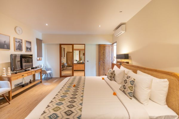 Cozy Balinese hotel bedroom with king bed, rattan headboard, ikat pillows, frangipani accents, TV, and wooden furniture on light floors.
