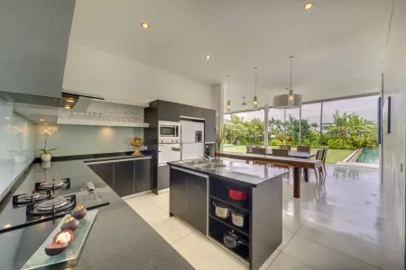 Modern open-plan kitchen with sleek black cabinets, central island, and tropical garden-pool view through expansive glass walls. - Bali Villas
