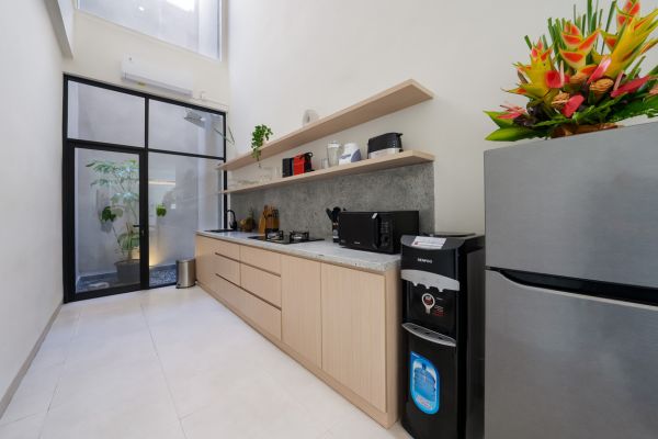 Modern minimalist kitchen with light wood cabinets, stainless steel fridge and appliances, open shelving, and a glass door to a tropical garden. - Bali Villas