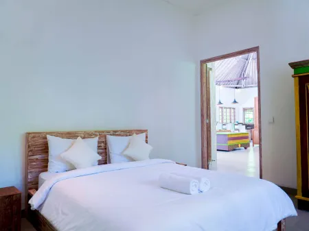 Cozy Balinese bedroom with carved wooden bed, crisp white linens, folded towel, colorful wardrobe, and open door to thatched-roof kitchen.
