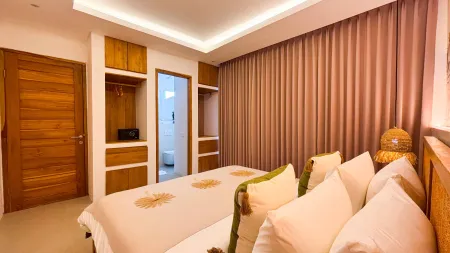Spacious tropical bedroom in a Balinese villa with wooden accents, king bed with beige linens and pillows, sheer curtains, and soft ambient lighting.