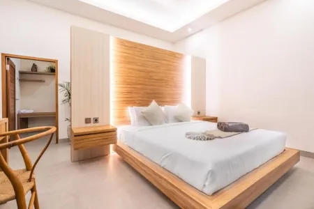 Modern minimalist bedroom featuring a low wooden platform bed with white linens, illuminated teak headboard, rattan chair, and potted plants against white walls. - Bali Villas